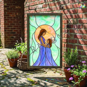 Può includere: Vetrata raffigurante una madre e un bambino nei toni del blu, marrone e arancione. L'opera è incorniciata in una cornice di legno scuro e appesa a un muro di mattoni. Piante in vaso e un cesto di vimini circondano l'immagine.
