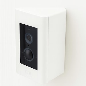 May include: A white, triangular-shaped security camera with a black rectangular face. The face features two circular lenses and the brand name "ring" at the bottom. The camera is mounted on a white wall.