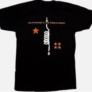 Może przedstawiać: Czarny t-shirt z pomarańczowym napisem "JOE STRUMMER & THE MESCALEROS". Biała grafika cewki ze strzałką i kroplą znajduje się pod tekstem. Dwie pomarańczowe gwiazdki po lewej i trzy po prawej.