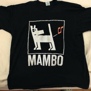 May include: Black t-shirt with a white graphic of a dog inside a white square, with a musical note and the word "MAMBO" in white block letters. A casual, unisex tee.