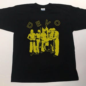 Op de afbeelding: Zwart T-shirt met het woord "DEVO" in gele blokletters boven een gele afbeelding van verschillende figuren. De figuren lijken bijpassende outfits en hoeden te dragen. Het shirt is van het merk Gildan.