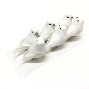 Puede incluir: Seis figuritas de palomas blancas con picos grises y ojos negros. Las palomas están dispuestas en fila sobre una superficie blanca.