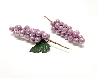 Puede incluir: Dos ramitas de uvas artificiales con una sola hoja verde en cada una. Las uvas son de color morado claro y tienen un acabado perlado.