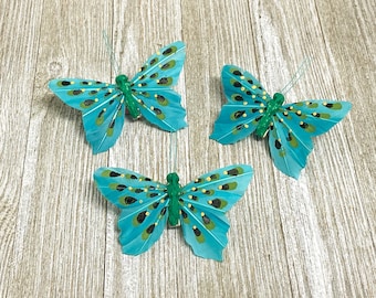 May include: Three teal blue butterfly decorations with green accents and black spots. The butterflies are made of feathers and have a wire stem.