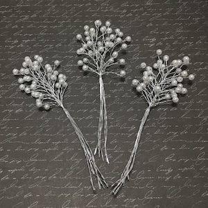 Puede incluir: Tres tallos florales de alambre plateado, cada uno adornado con racimos de perlas blancas. Los tallos están dispuestos sobre un fondo oscuro con un sutil patrón de escritura, creando una estética elegante y delicada. Ideal para manualidades o decoración de bodas.