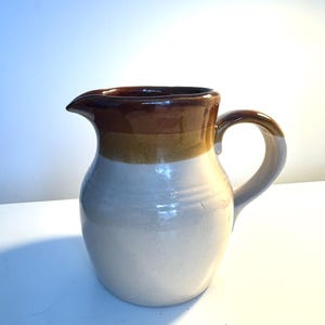 May include: A ceramic pitcher with a rounded body and a handle. The pitcher has a white base, with a band of yellow and brown near the top, and a dark brown rim and spout. The pitcher is designed for serving beverages.