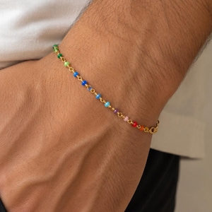 Könnte beinhalten: Ein zartes Armband aus einer Goldkette mit einer Reihe kleiner, runder, mehrfarbiger Perlen in den Farben Grün, Blau, Violett, Rosa und Rot. Das Armband besteht aus einer feinen Metallgliederkette und einem kleinen, runden Verschluss.