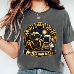 May include: Dark gray t-shirt featuring a circular graphic with the words "AMAZE!" and "PROJECT HAIL MARY." The graphic depicts an astronaut and a robotic spider-like creature in a space scene. The shirt is a comfortable, casual style.