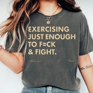 May include: Dark gray t-shirt with the text "EXERCISING JUST ENOUGH TO F≈CK & FIGHT." in a tan color. The shirt also has the text "Not for posies" and "Trend" in a smaller font.