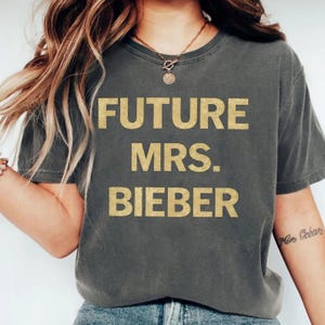 May include: Dark gray t-shirt with the text "FUTURE MRS. BIEBER" in gold glitter lettering. The shirt has a crew neck and short sleeves. The model is wearing a gold necklace.