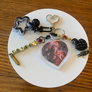 May include: A collection of keychains with various charms. Includes a star-shaped charm, a heart-shaped charm, and a heart-shaped pendant with a character image. The word "SYLUS" is spelled out on a beaded strand.