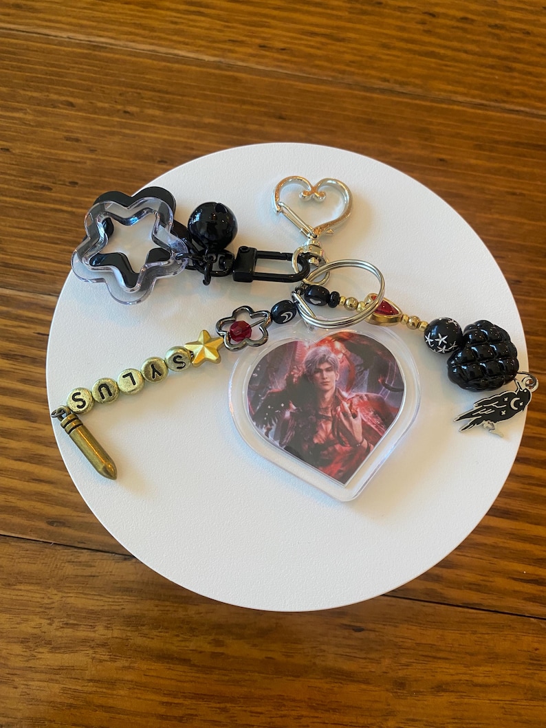 May include: A collection of keychains with various charms. Includes a star-shaped charm, a heart-shaped charm, and a heart-shaped pendant with a character image. The word "SYLUS" is spelled out on a beaded strand.