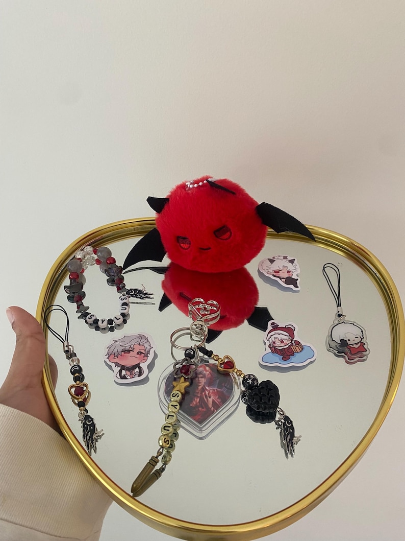 May include: A collection of accessories, including a red plush devil, keychains, and stickers, arranged on a heart-shaped mirror tray with a gold rim. The keychains feature beads, charms, and the word "stylus".
