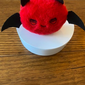 May include: A plush red devil with black horns and wings. The devil has embroidered eyes and a small smile. The plush is sitting on a white circular base. The background is a wooden surface.
