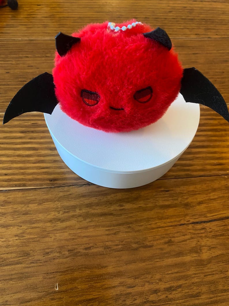 May include: A plush red devil with black horns and wings. The devil has embroidered eyes and a small smile. The plush is sitting on a white circular base. The background is a wooden surface.