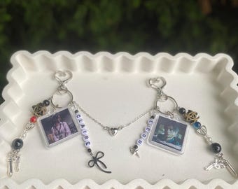 May include: Two keyrings linked by a chain with a heart charm. Each keyring features a photo in a clear square, with the names "CLAIRE" and "LEON" spelled out in beads. Decorative charms include a bow, a gun, and beads.