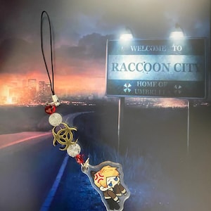 May include: A black lanyard with a charm featuring a cartoon character with an angry expression. The charm is attached to a string of red and clear beads, and a gold biohazard symbol. The background shows a sign that reads "Welcome to Raccoon City."