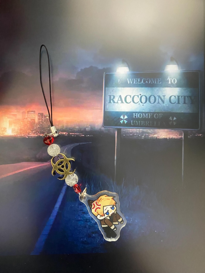 May include: A black lanyard with a charm featuring a cartoon character with an angry expression. The charm is attached to a string of red and clear beads, and a gold biohazard symbol. The background shows a sign that reads "Welcome to Raccoon City."