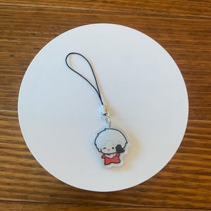 May include: A clear acrylic charm featuring a cartoon character with white hair and a red outfit. The charm has a black string attached for hanging. The charm is on a white circular surface.
