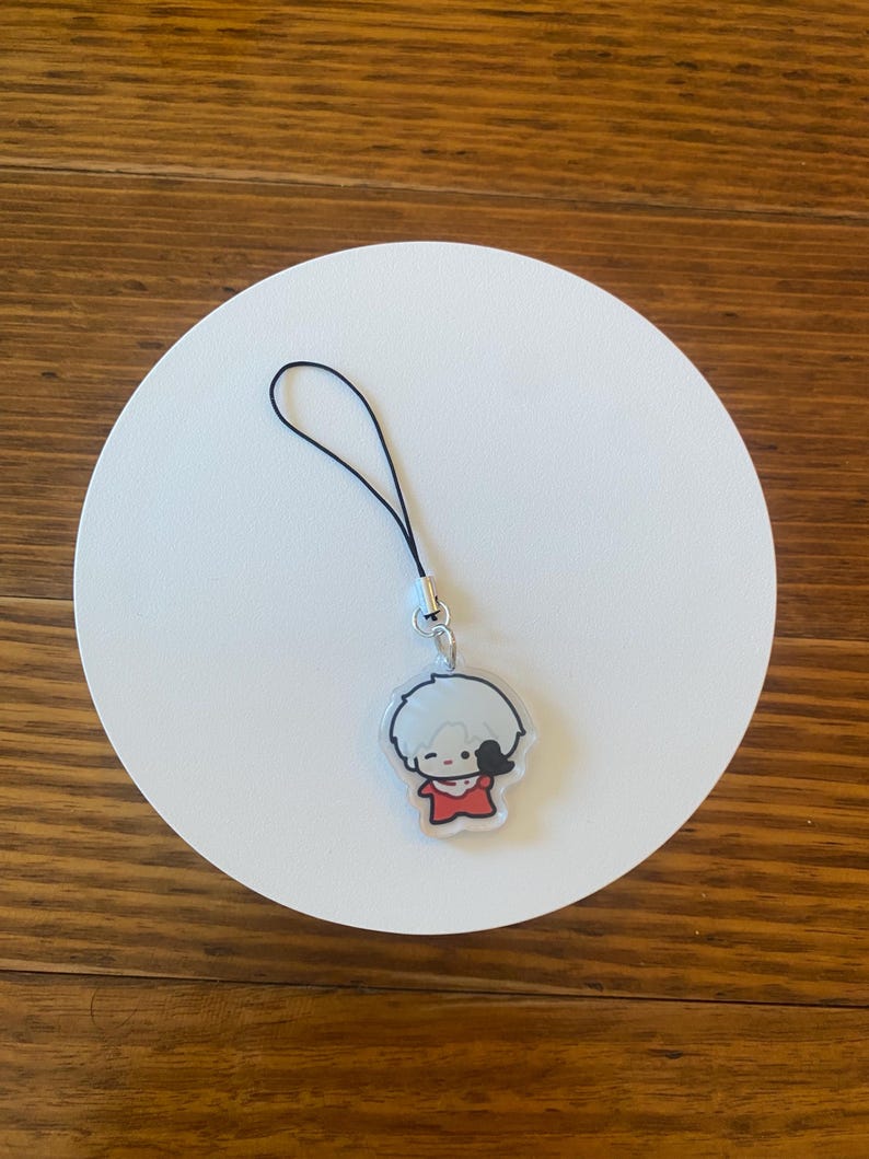 May include: A clear acrylic charm featuring a cartoon character with white hair and a red outfit. The charm has a black string attached for hanging. The charm is on a white circular surface.
