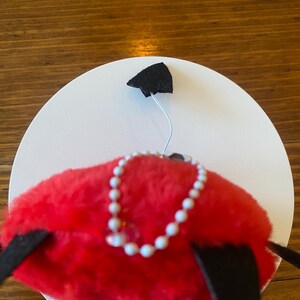 May include: A whimsical red plush hat with a white pearl necklace and black accents. The hat is set against a white circular backdrop, with a black triangular shape attached to the top.