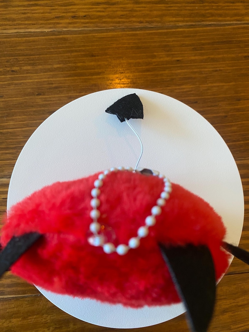 May include: A whimsical red plush hat with a white pearl necklace and black accents. The hat is set against a white circular backdrop, with a black triangular shape attached to the top.