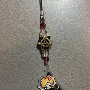 May include: A black lanyard with a charm featuring a cartoon character with blonde hair and an angry expression. The charm is attached to a string of red and clear beads, and a gold-colored metal charm.