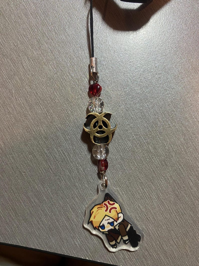 May include: A black lanyard with a charm featuring a cartoon character with blonde hair and an angry expression. The charm is attached to a string of red and clear beads, and a gold-colored metal charm.