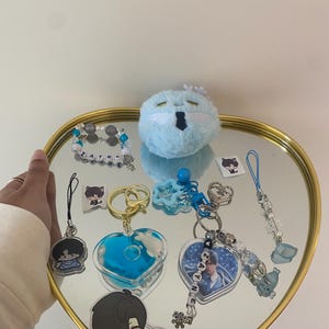 May include: A collection of keychains, charms, and a plushie arranged on a heart-shaped mirror tray with a gold rim. The items include heart-shaped and circular keychains with blue and white accents, and a blue plushie.