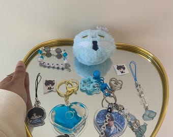 Op de afbeelding: Een verzameling sleutelhangers, bedels en een pluche speelgoed op een hartvormige spiegellade met een gouden rand. De items omvatten hartvormige en ronde sleutelhangers met blauwe en witte accenten, en een blauw pluche speelgoed.