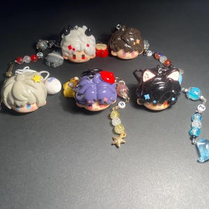 May include: A collection of six character-themed beaded charms. The charms feature various hair colors and accessories, including horns, stars, and cat ears. Each charm is connected to a beaded strand with decorative elements.