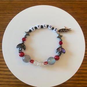 May include: A beaded bracelet with the word "SYLUS" spelled out in white letter beads. The bracelet features red, gray, and clear beads, along with star and mushroom charms. A gold-colored charm is also attached.