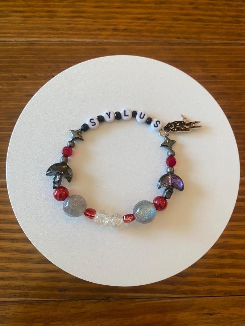 May include: A beaded bracelet with the word "SYLUS" spelled out in white letter beads. The bracelet features red, gray, and clear beads, along with star and mushroom charms. A gold-colored charm is also attached.
