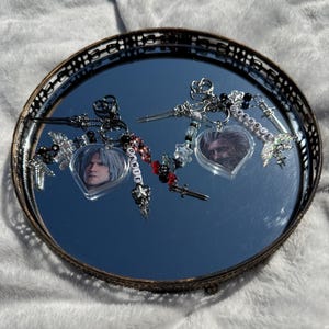 May include: Two heart-shaped keyrings featuring images of a person with white hair. The keyrings have silver charms, beads, and the names "Dante" and "Vergil" spelled out in beads. They are displayed on a round, mirrored tray.