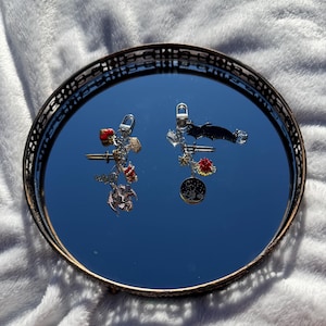 Puede incluir: Dos llaveros con dijes, exhibidos en un espejo redondo con borde dorado. Un llavero presenta un dragón, rosas rojas y una espada. El otro tiene un murciélago, una luna y estrellas, y una rosa roja. El espejo está sobre una superficie blanca y esponjosa.