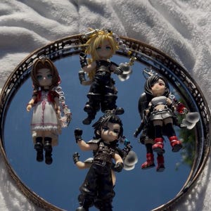 Puede incluir: Cuatro figuras de personajes de Final Fantasy se exhiben en una bandeja redonda y espejada con borde decorativo. Las figuras están en varias poses y atuendos, con detalles y colores vibrantes. La bandeja se encuentra sobre una superficie blanca y esponjosa.