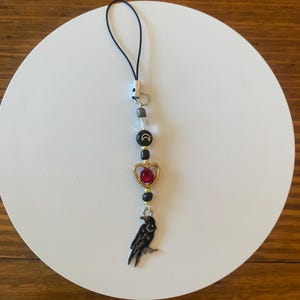 May include: A black and gold charm with a raven pendant, red heart, and moon and star beads. The charm is strung on a black cord and features a variety of beads in clear, black, and gold colors. The charm is designed to be attached to a phone or other small item.