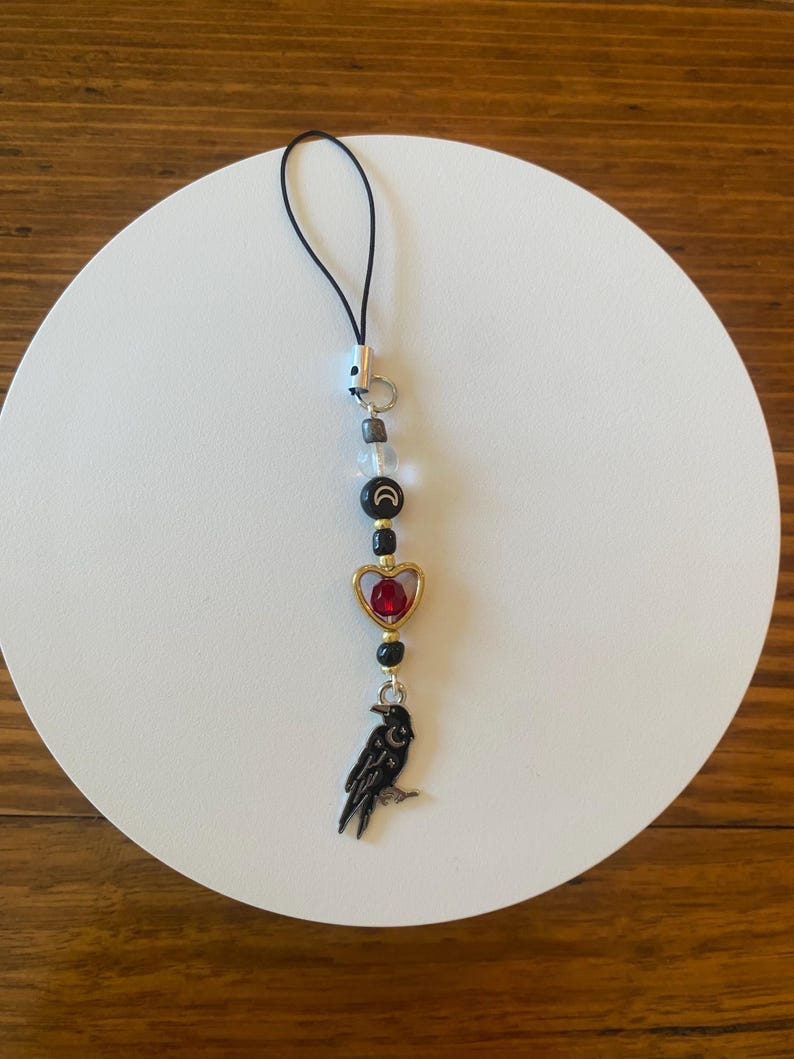 May include: A black and gold charm with a raven pendant, red heart, and moon and star beads. The charm is strung on a black cord and features a variety of beads in clear, black, and gold colors. The charm is designed to be attached to a phone or other small item.