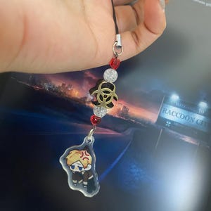 May include: A black lanyard with a charm featuring a cartoon character with blonde hair and an angry expression. The charm is attached to a string of red and clear beads, and a gold-colored biohazard symbol.