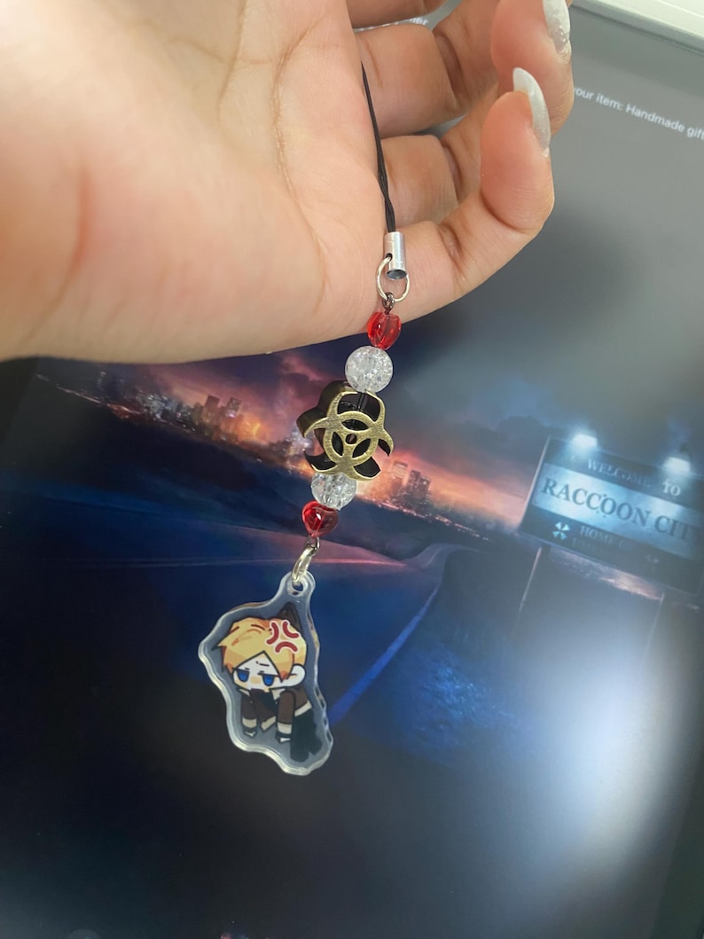 May include: A black lanyard with a charm featuring a cartoon character with blonde hair and an angry expression. The charm is attached to a string of red and clear beads, and a gold-colored biohazard symbol.