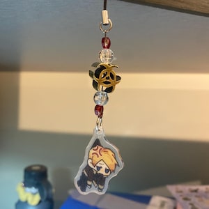 May include: A black lanyard with a charm featuring a cartoon character with blonde hair and an angry expression. The charm is attached to a decorative piece with clear beads, red beads, and a gold and black flower.
