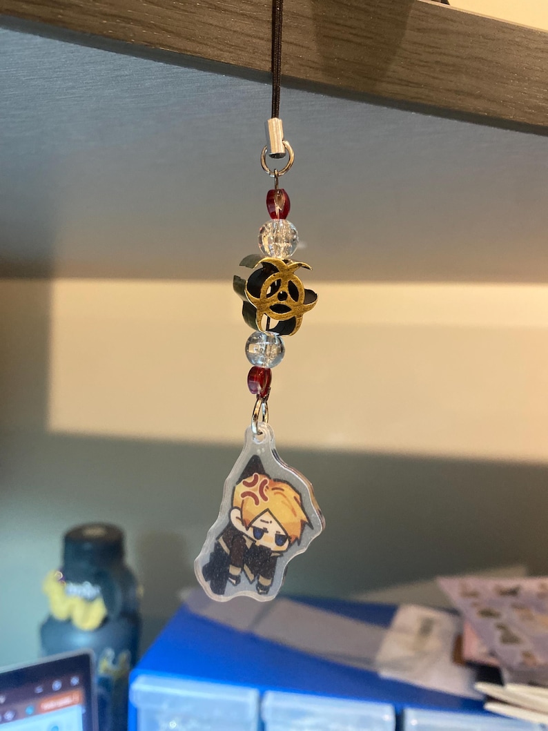 May include: A black lanyard with a charm featuring a cartoon character with blonde hair and an angry expression. The charm is attached to a decorative piece with clear beads, red beads, and a gold and black flower.