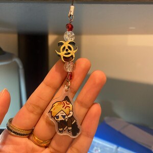 May include: A clear acrylic charm with a cartoon character, featuring a gold biohazard symbol and red and clear beads. The charm is attached to a black string.