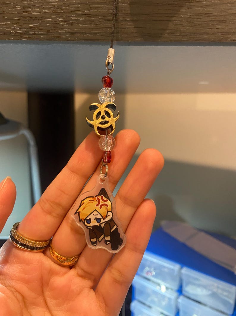 May include: A clear acrylic charm with a cartoon character, featuring a gold biohazard symbol and red and clear beads. The charm is attached to a black string.