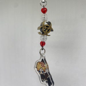 May include: A beaded charm with a black cord, featuring red and clear beads, a gold-colored biohazard symbol, and a clear acrylic charm with a cartoon character illustration. The character has blonde hair and is wearing a brown outfit.