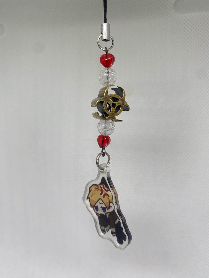 May include: A beaded charm with a black cord, featuring red and clear beads, a gold-colored biohazard symbol, and a clear acrylic charm with a cartoon character illustration. The character has blonde hair and is wearing a brown outfit.