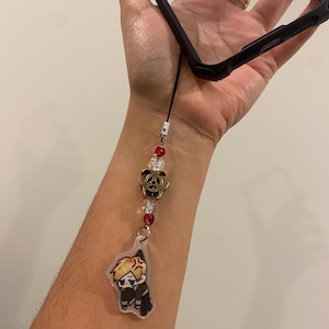 May include: Black phone case with a decorative charm. The charm features a cartoon character, red and clear beads, and a gold-colored rose. The charm is attached to the phone case with a black cord.