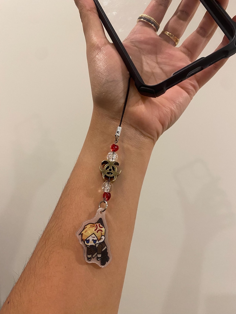 May include: Black phone case with a decorative charm. The charm features a cartoon character, red and clear beads, and a gold-colored rose. The charm is attached to the phone case with a black cord.