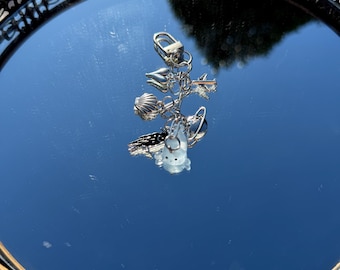 May include: A silver keychain with multiple charms, including a shell, a plane, a rocket, and a small, translucent bear. The charms are attached to a silver clasp. The keychain is displayed on a mirrored tray with a decorative edge.