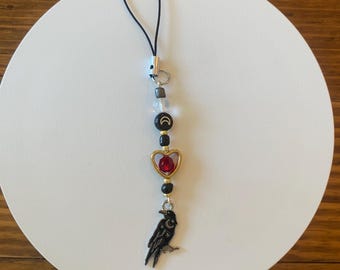 May include: A black and gold charm with a raven pendant, red heart, and moon and star beads. The charm is strung on a black cord and features a variety of beads in clear, black, and gold colors. The charm is designed to be attached to a phone or other small item.